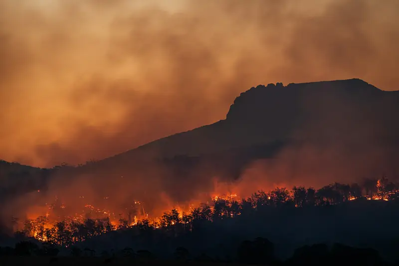 Wildfire burning along a hillside at dusk under heavy smoke